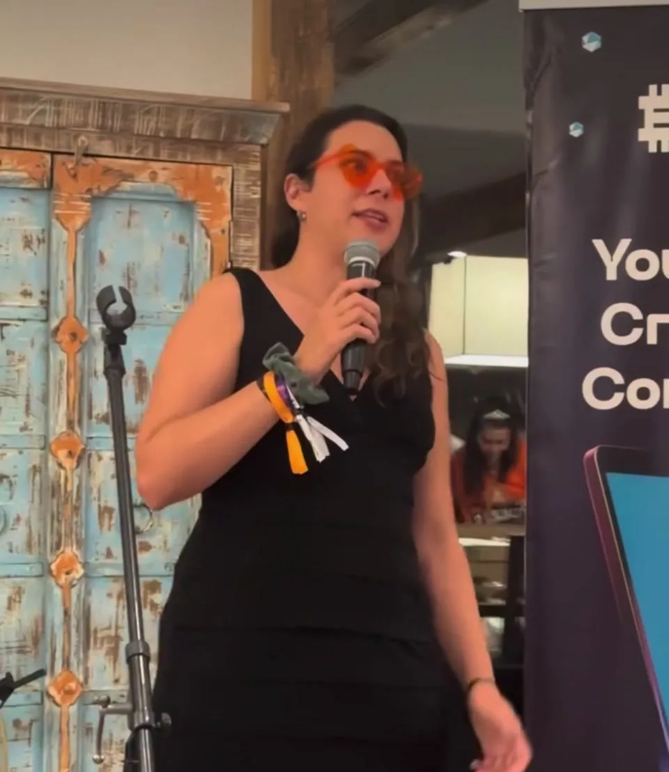 Steph Ferrera speaking into a handheld microphone at BitBasel, in orange glasses, a crypto conference banner behind her.